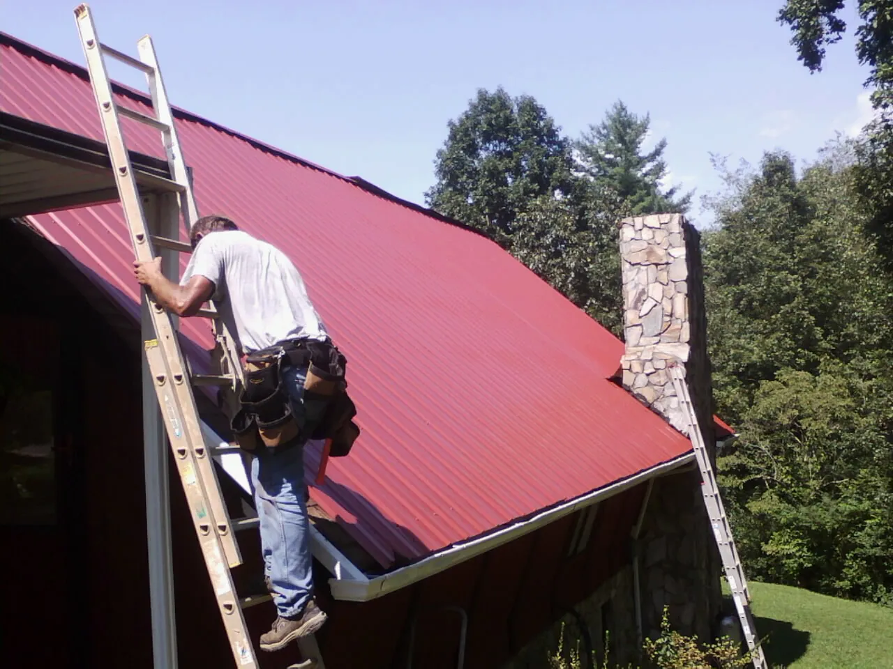 Roofer on a ladder beside a red metal roof and stone chimney in Doerun